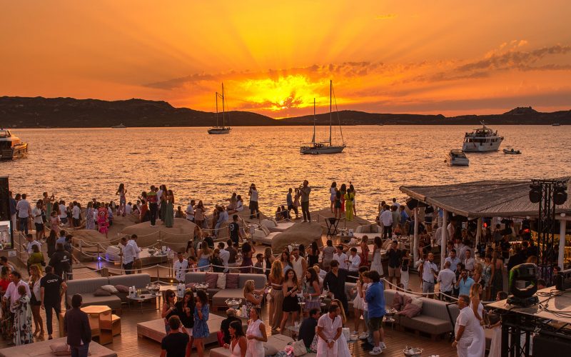 Sunset by the sea with boats in the background and people at Phi Beach club in Baja Sardinia, Italy