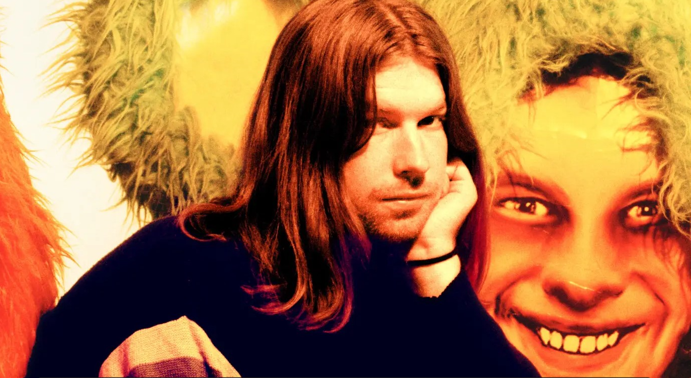 A man (Aphex Twin) with long hair in front of a painting