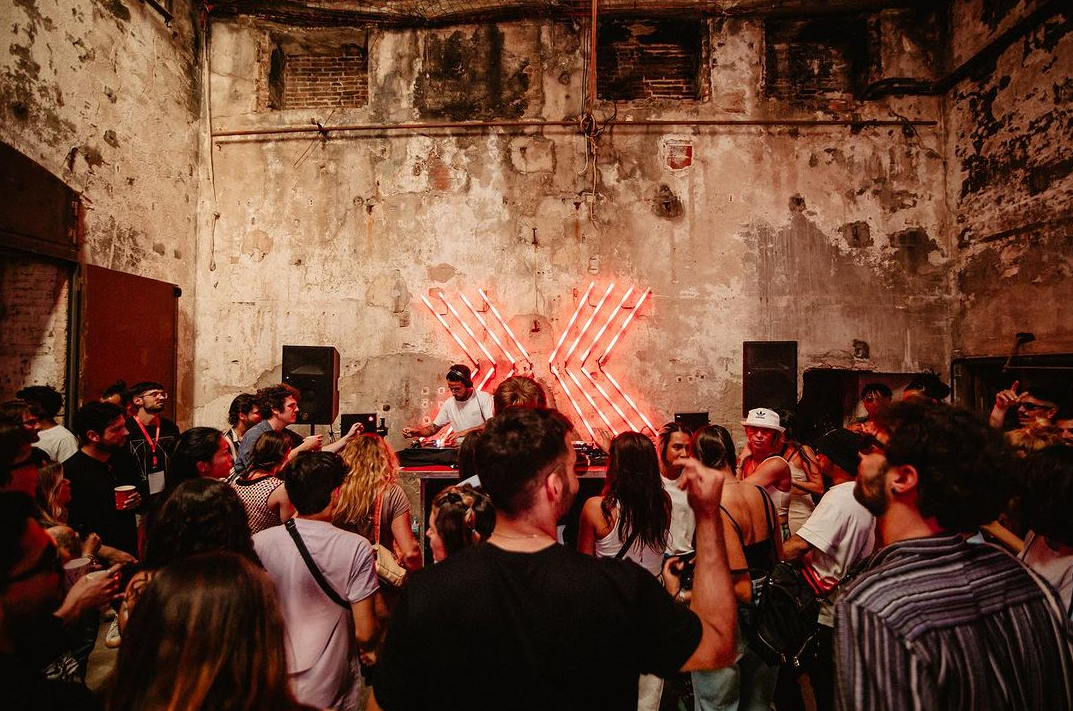 crowd of people dancing in front of a dj in a warehouse at bridge festival