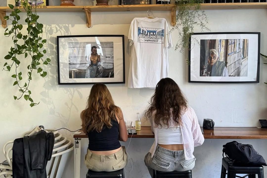 Two women sitting at a bar-height counter in Neighborhood Café, Lisbon, with wine bottles, framed photos, and hanging plants decorating the wall