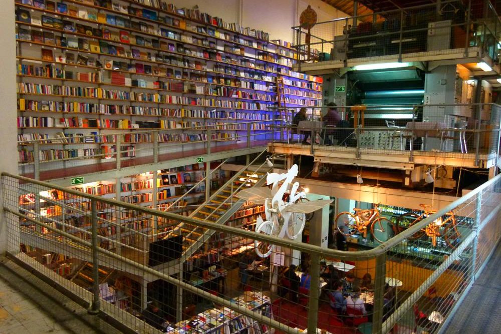 Interior of Ler Devagar bookstore in Lisbon, featuring towering bookshelves, an old printing press, and a suspended bicycle sculpture.