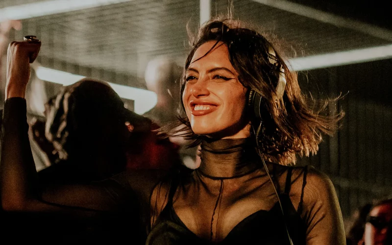 Sara Landry smiling mid-set with headphones on, wearing a sheer black outfit as the crowd dances behind her.