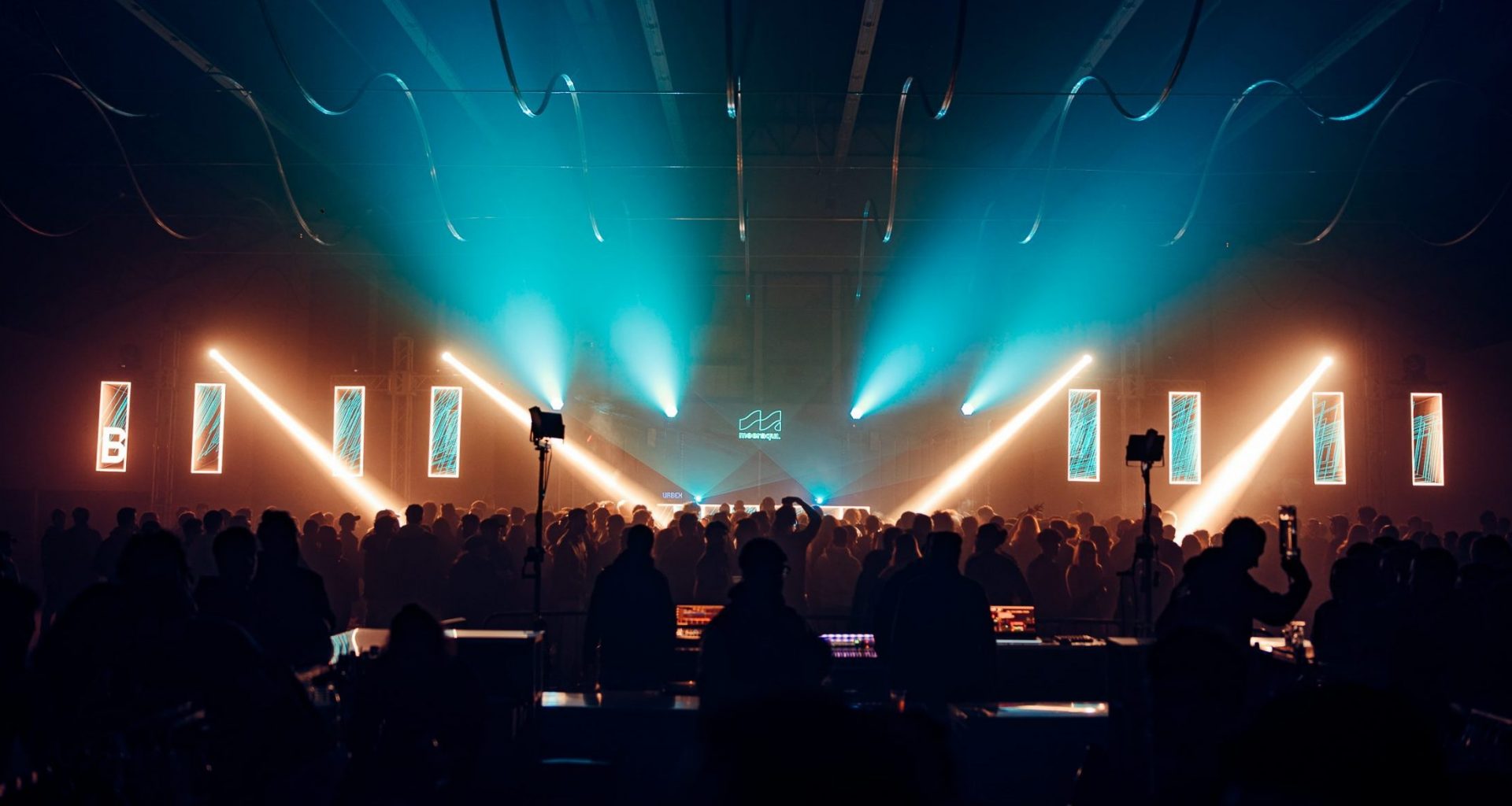 Vibrant crowd gathered at Meeraqui event in a historic venue, illuminated by colorful lights and lasers during an electronic music performance.