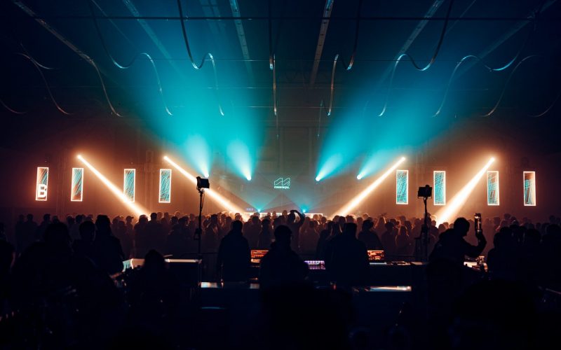 Vibrant crowd gathered at Meeraqui event in a historic venue, illuminated by colorful lights and lasers during an electronic music performance.