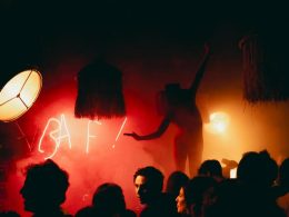 Performer silhouetted against red smoke and neon BAF! sign during a crowded late-night party, surrounded by eclectic lighting and decor