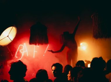 Performer silhouetted against red smoke and neon BAF! sign during a crowded late-night party, surrounded by eclectic lighting and decor