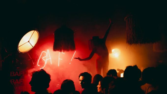 Performer silhouetted against red smoke and neon BAF! sign during a crowded late-night party, surrounded by eclectic lighting and decor