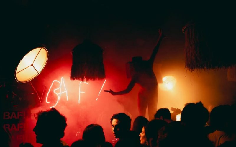 Performer silhouetted against red smoke and neon BAF! sign during a crowded late-night party, surrounded by eclectic lighting and decor