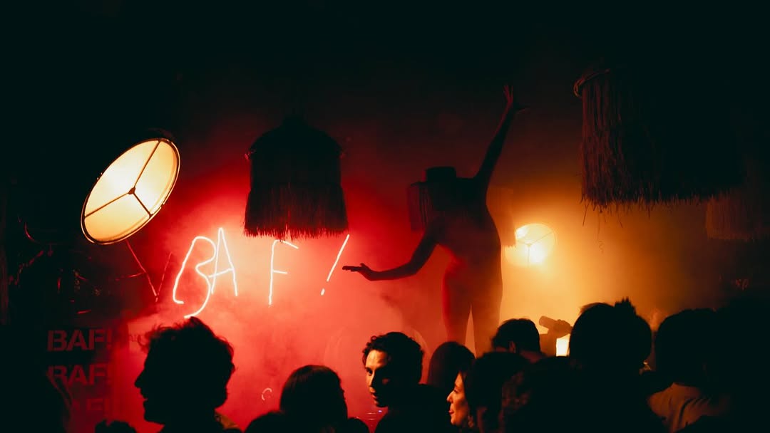 Performer silhouetted against red smoke and neon BAF! sign during a crowded late-night party, surrounded by eclectic lighting and decor