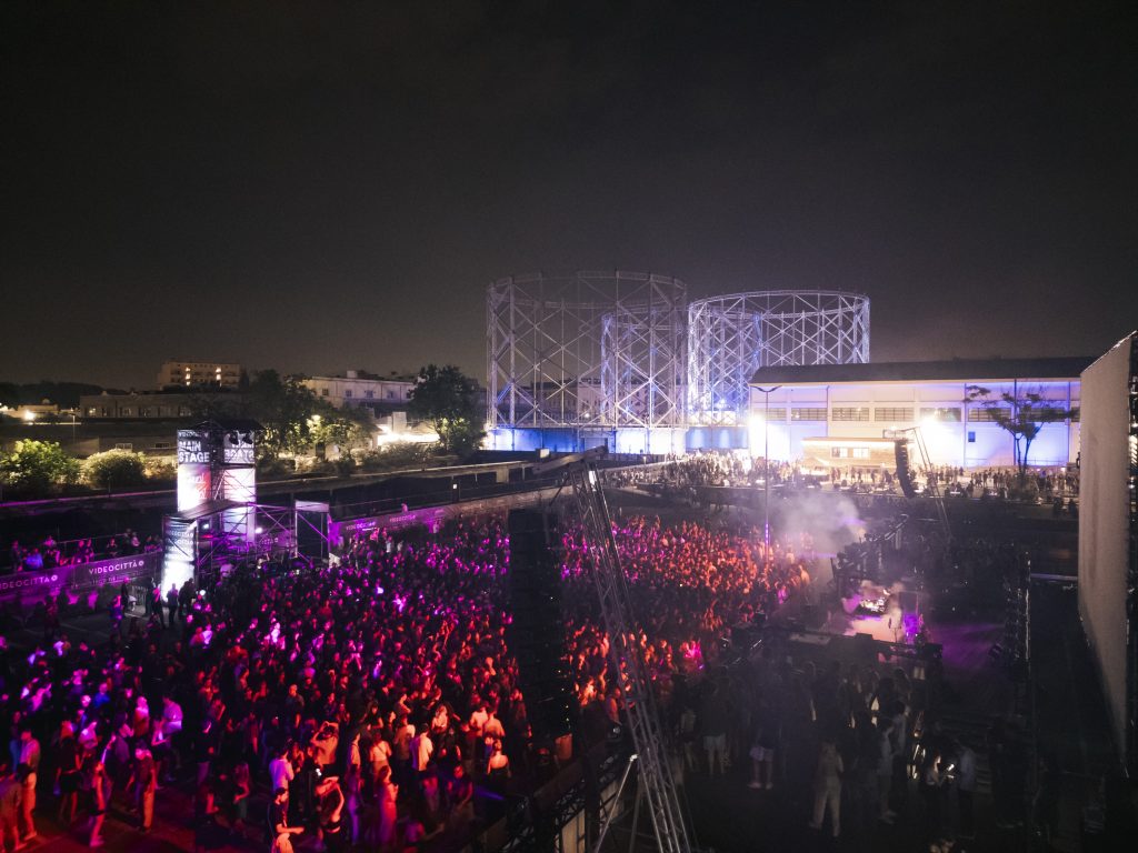 Night crowd shot of VIDEOCITTÀ Festival under the illuminated Rome gasometer structure.