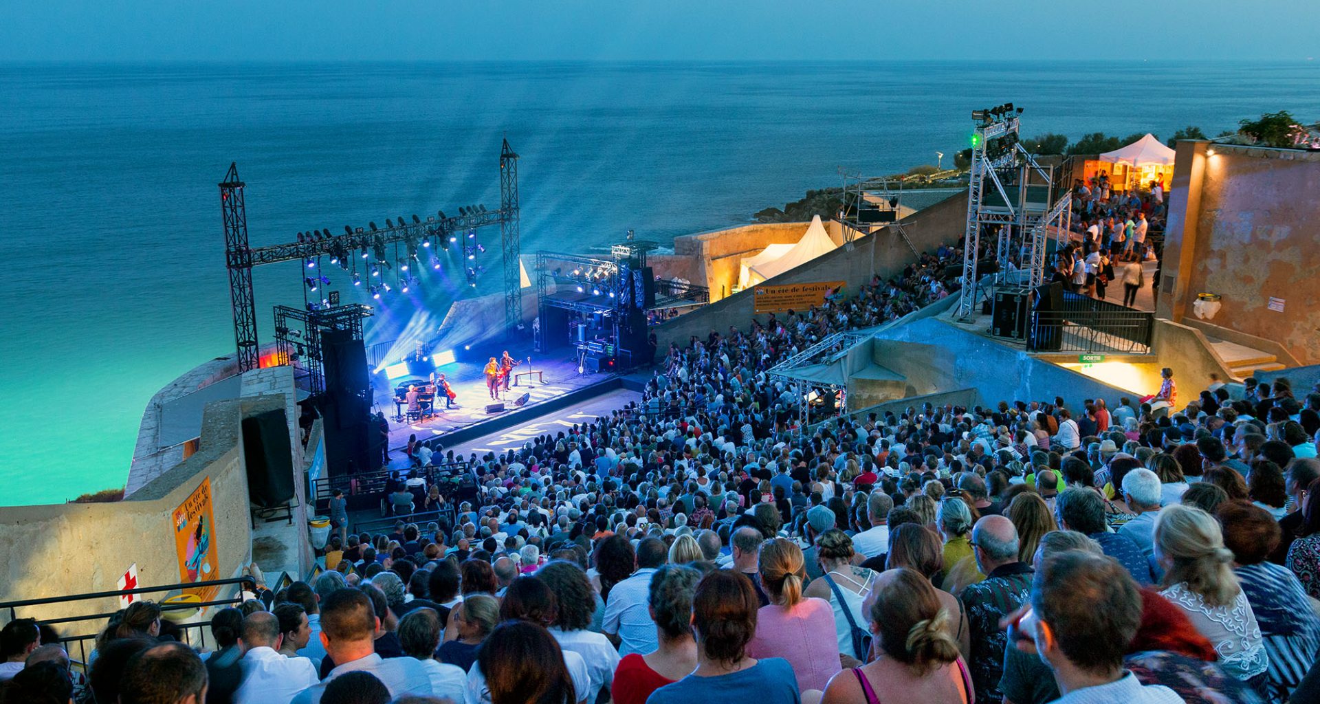A packed open-air concert at Théâtre de la Mer in Sète, overlooking the Mediterranean, with stage lights beaming into the night sky.
