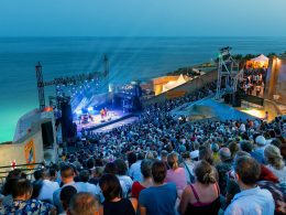 A packed open-air concert at Théâtre de la Mer in Sète, overlooking the Mediterranean, with stage lights beaming into the night sky.