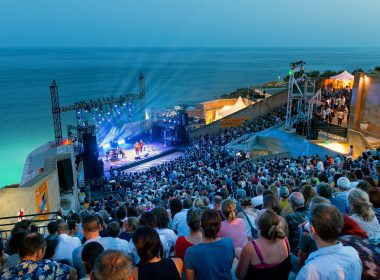 A packed open-air concert at Théâtre de la Mer in Sète, overlooking the Mediterranean, with stage lights beaming into the night sky.