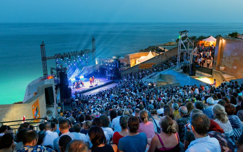 A packed open-air concert at Théâtre de la Mer in Sète, overlooking the Mediterranean, with stage lights beaming into the night sky.