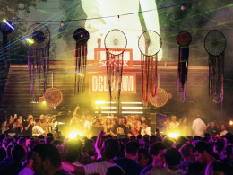 Delirium XS crowd shot — A vibrant crowd dancing under dreamcatcher-style decorations and colorful lights at Delirium XS, with DJs performing on the Open Air Stage
