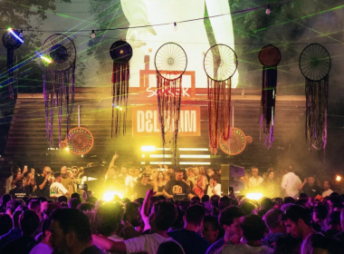 Delirium XS crowd shot — A vibrant crowd dancing under dreamcatcher-style decorations and colorful lights at Delirium XS, with DJs performing on the Open Air Stage