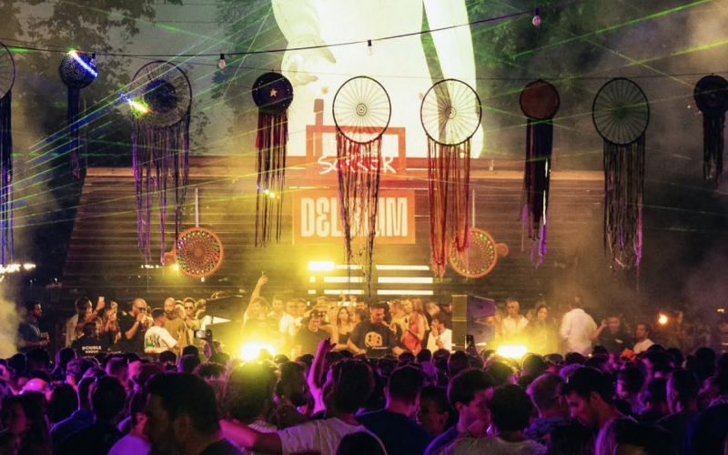Delirium XS crowd shot — A vibrant crowd dancing under dreamcatcher-style decorations and colorful lights at Delirium XS, with DJs performing on the Open Air Stage