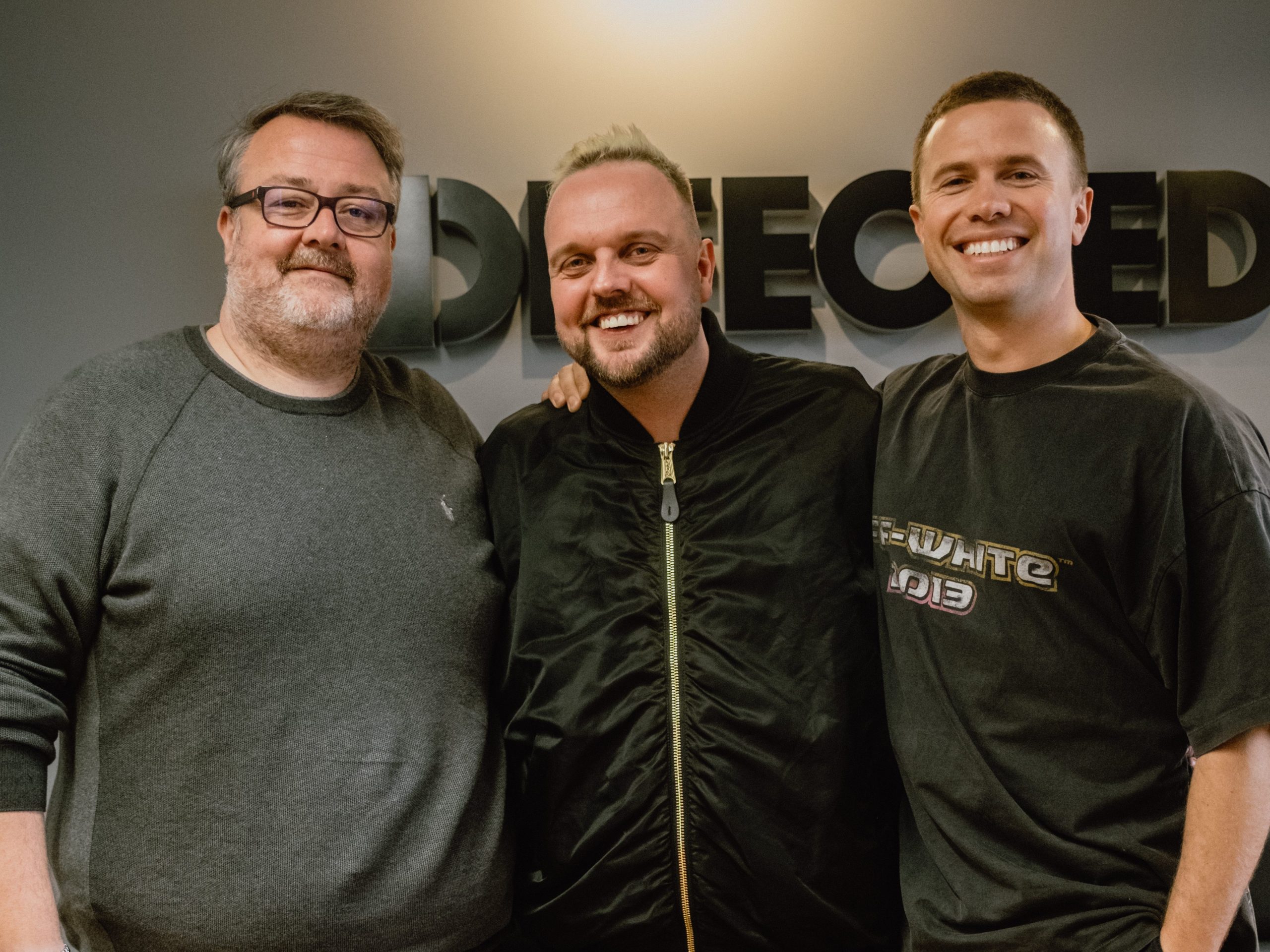 Wez Saunders, Steven Braines, and George Pritchard posing in front of the Defected sign
