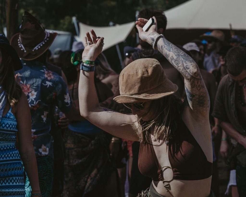 Tattooed festival-goer in a brown bucket hat dances with hands raised at a Beyond The Mountain event, crowd and tents in the background.