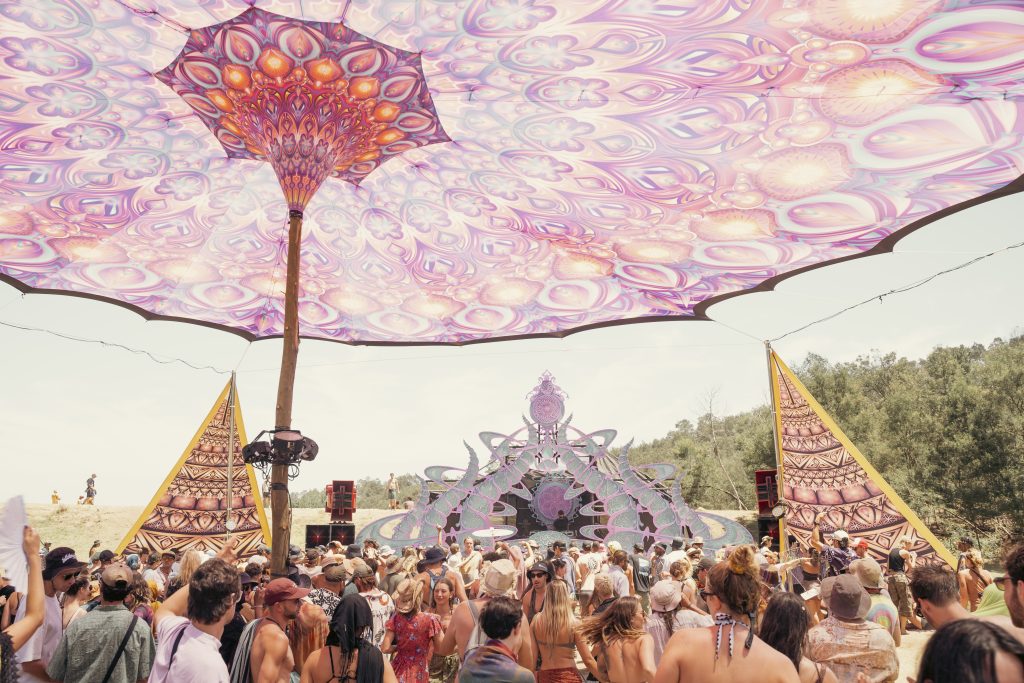 Beyond The Mountain festival with a crowd dancing under a large psychedelic pink canopy, facing a geometric art stage in a forest clearing.