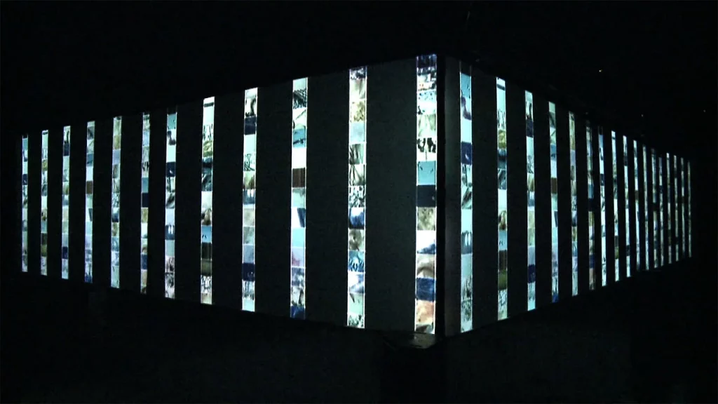 Dark-room audiovisual installation: a rectangular structure wrapped in vertical light strips projecting fragmented nature footage — by Ryoichi Kurokawa.