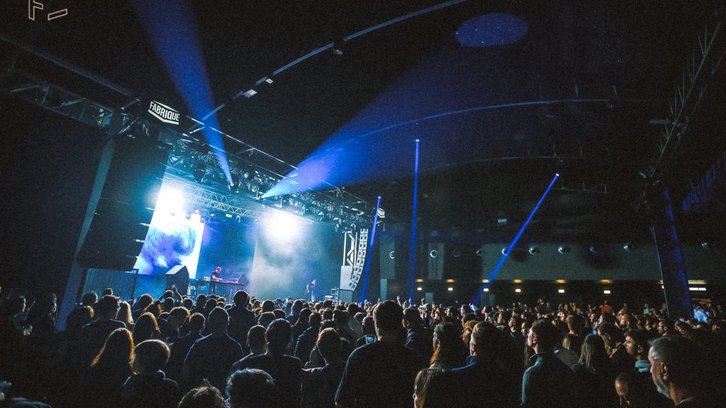 Foule compacte à FABRIQUE devant un DJ sur scène, faisceaux bleus balayant la salle sombre avec un écran LED lumineux derrière.