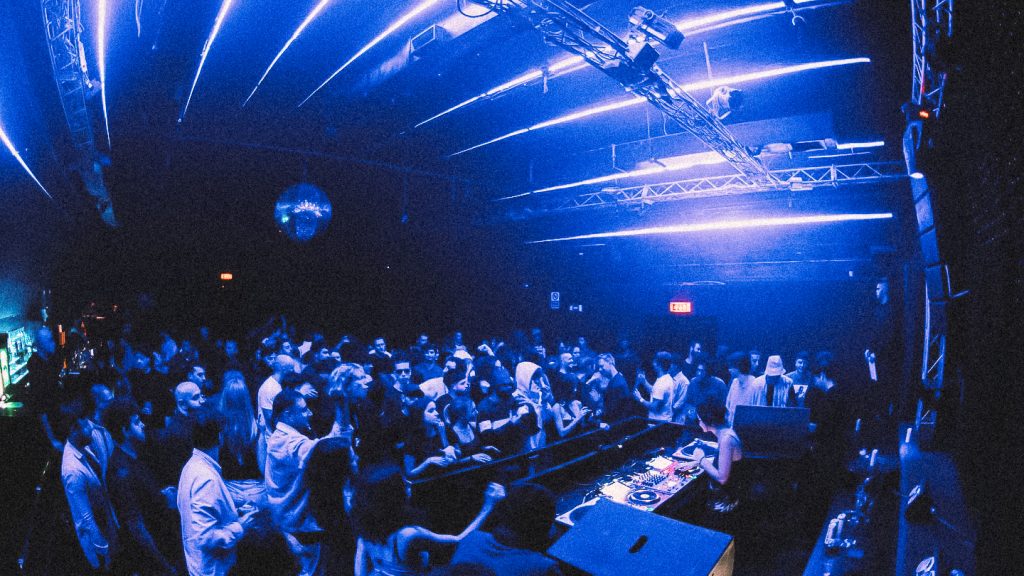 Tunnel Club bondé sous lumières bleues, foule serrée face au DJ booth, néons au plafond et boule à facettes en arrière-plan.