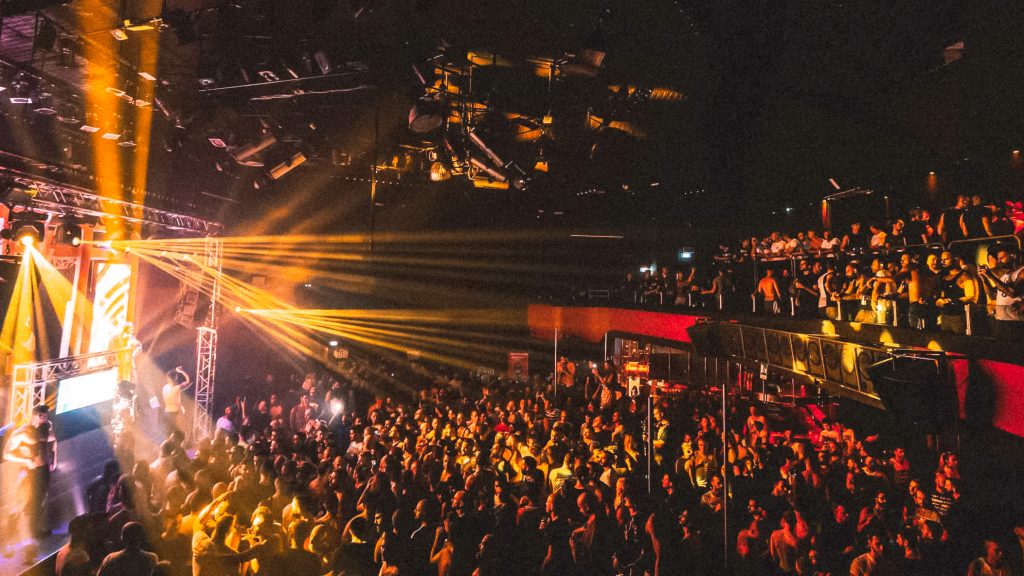 Foule compacte au District 272, faisceaux lumineux dorés balayant la scène et les balcons d’un grand club à plusieurs niveaux.