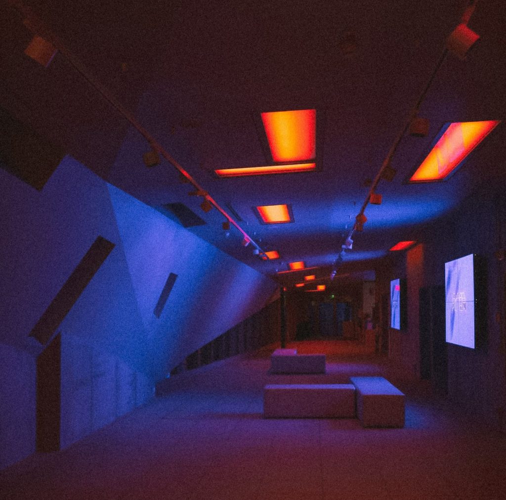 Couloir futuriste de l’ARCA Milano, éclairé de panneaux rouges et bleus, avec murs géométriques et bancs minimalistes.
