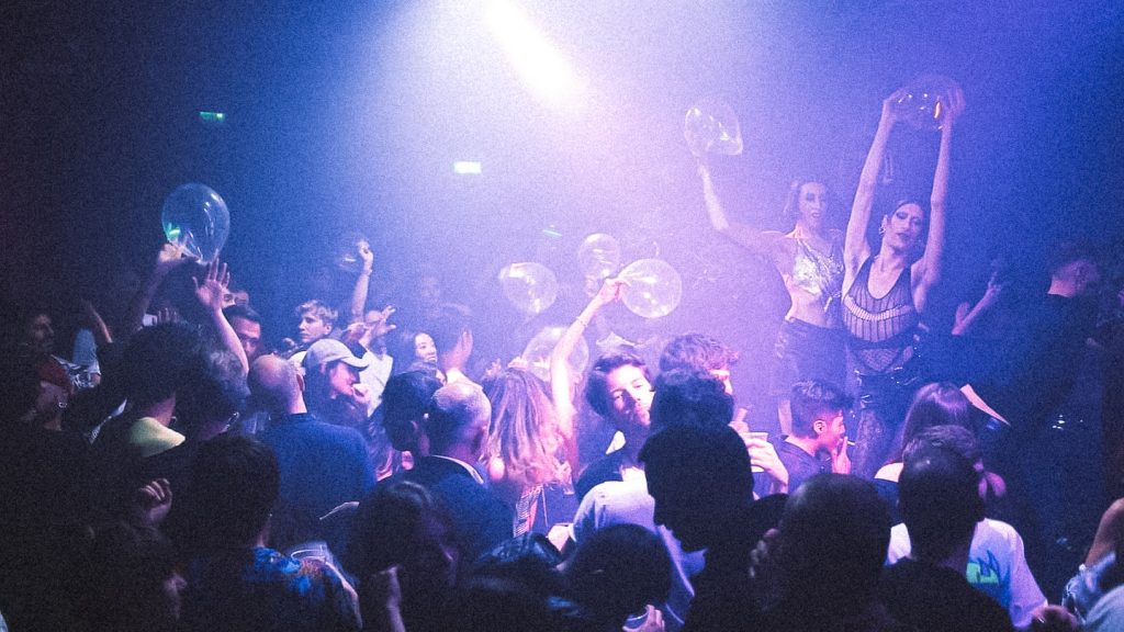 Soirée bondée au Q Club, piste enfumée sous lumières violettes avec danseuses surélevées brandissant des ballons transparents au-dessus de la foule.