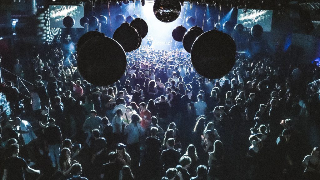 Foule dense à Alcatraz (Milan), grande piste de danse sous boules suspendues et boule disco, scène vivement éclairée au fond.
