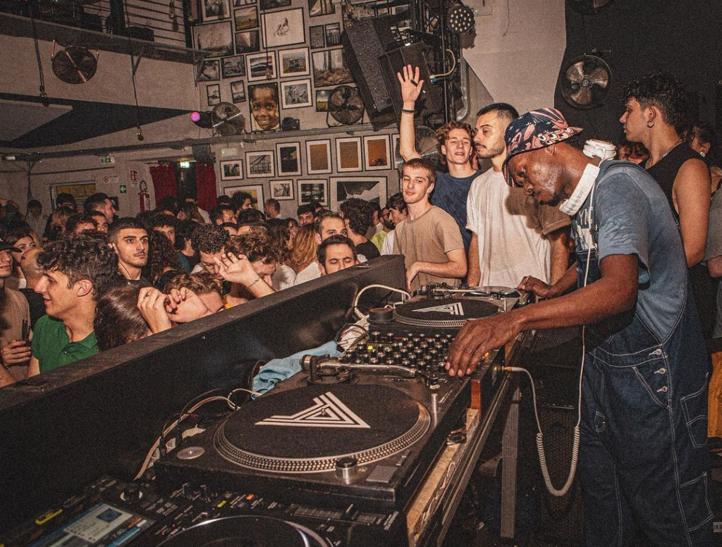 Packed crowd at Club Alcazar in Rome gathered around the DJ booth, turntables in the foreground and walls covered with framed art.