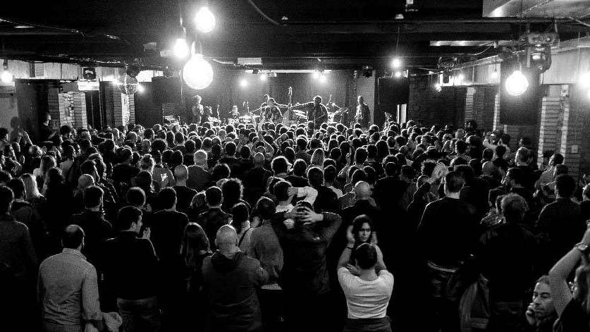 Crowd shot at Monk Club in Rome, audience facing a distant stage with hanging bulbs illuminating the room.
