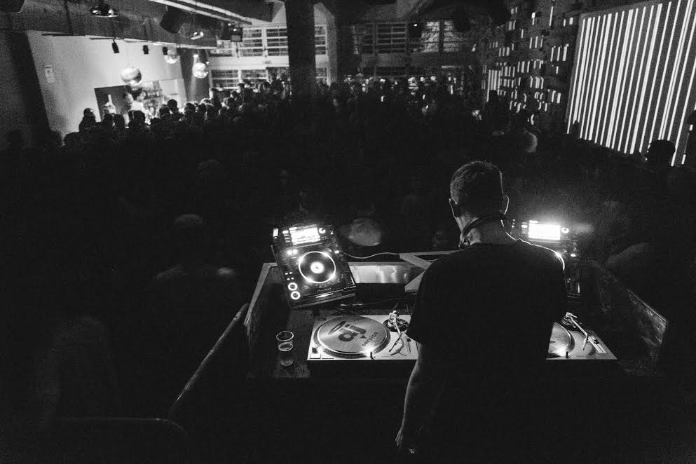 Black-and-white view from behind the DJ at Lanificio 159 in Rome, packed dance floor and a striped light wall to the right.