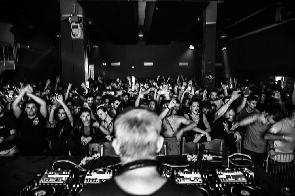 Black-and-white view from the DJ booth at Cieloterra, packed crowd with hands raised and decks in the foreground.