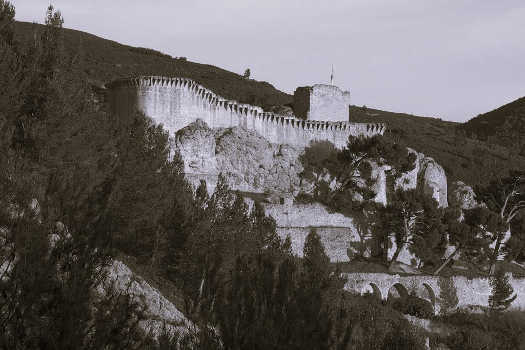 Black-and-white view of Fort de l’Antenne: long crenellated stone walls and a square tower running along a rocky hillside framed by trees.