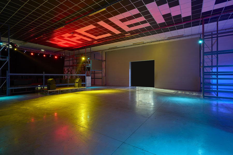 Empty warehouse-style hall at Hacienda, grid ceiling spelling “HACIENDA,” scaffolding along the walls, and colorful floor lights around a small side stage.