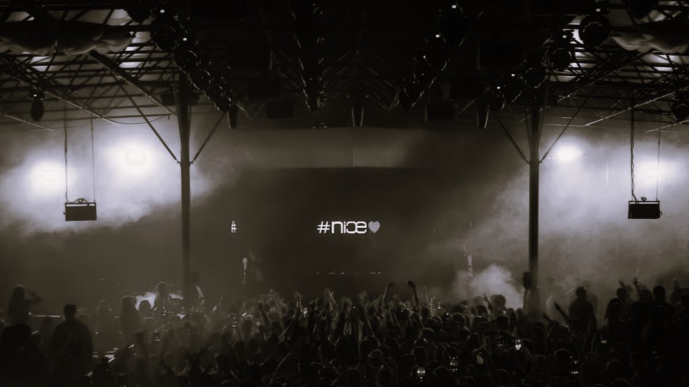 Black-and-white crowd at Nice Disco, hands raised under an industrial ceiling, haze and a backlit stage displaying “#nice”.