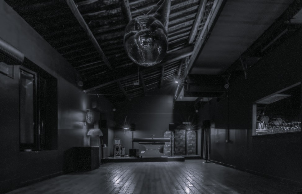 Empty black-and-white interior of Circolo dei Cerchi: wooden floor, sloped rafters with a hanging disco ball, small stage and a side bar.