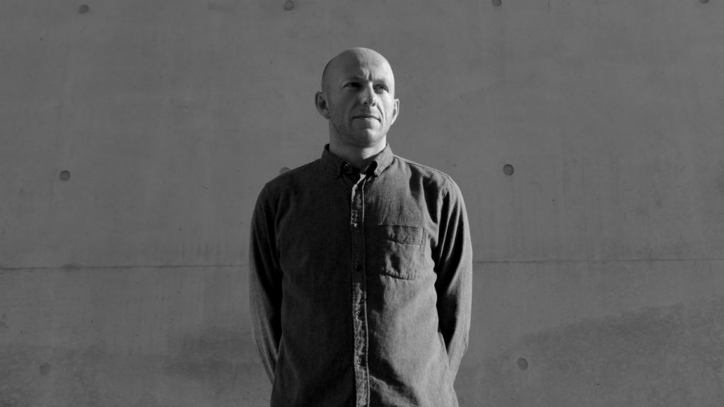 A black and white image of the DJ DVS1 standing in front of building.