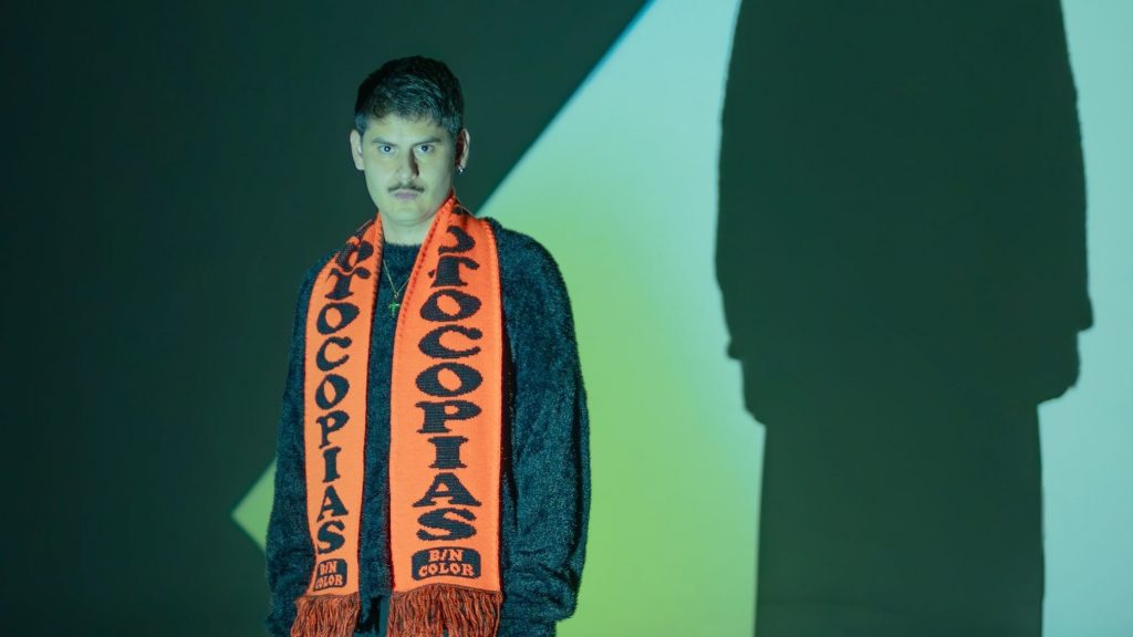 Foto del DJ Verraco con una bufanda naranja delante de un proyector de una pantalla verde 