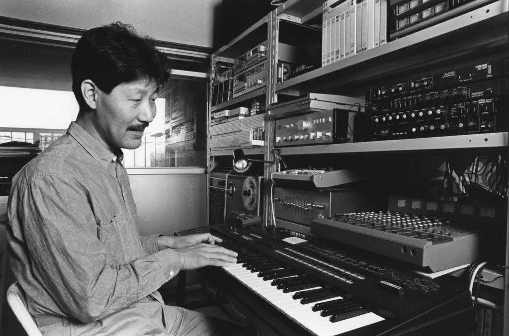 Black-and-white photo of Hiroshi Yoshimura playing a keyboard in his home studio, surrounded by vintage synthesizers, tape machines, and audio equipment used for ambient music production.