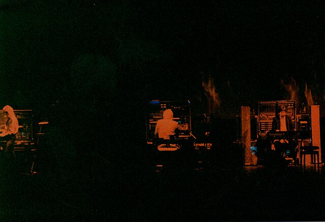 Dark, atmospheric live electronic performance, with three musicians surrounded by synthesizers and analog equipment, lit by minimal warm lighting against a deep black background.