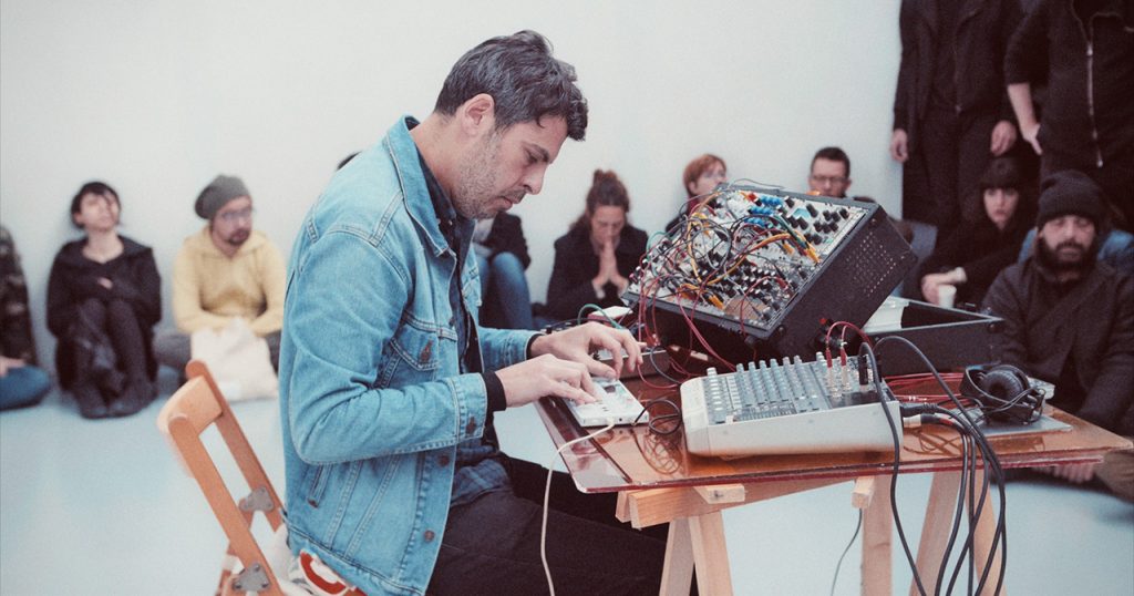 Live electronic performance in an intimate setting, with a musician seated at a table manipulating modular synthesizers and mixing equipment, surrounded by a quiet, attentive audience.