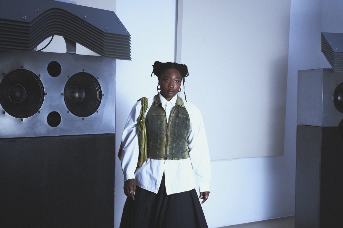 A woman named Kokoprisci posing in a white room in front of a hifi sound system