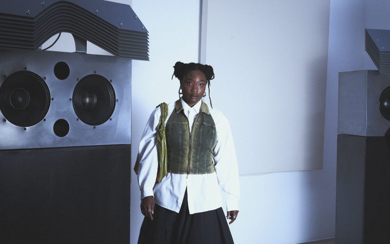 A woman named Kokoprisci posing in a white room in front of a hifi sound system